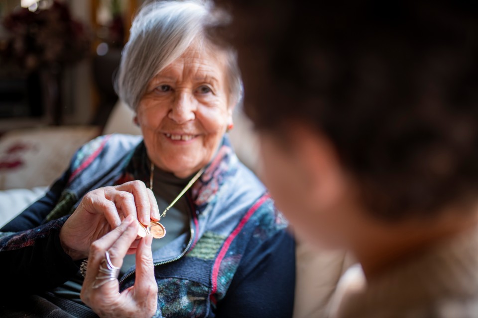 A smiling elderly woman holds a necklace coin between her fingers